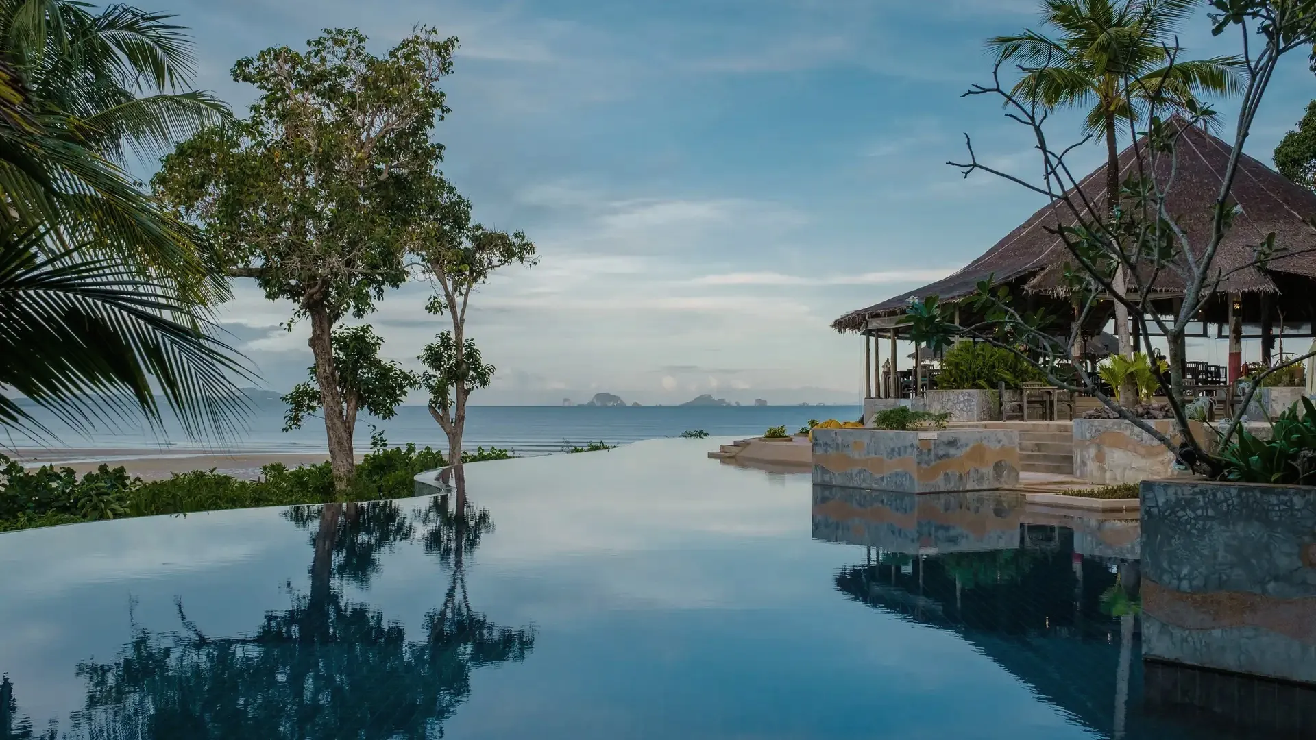 Infinity pool reflecting trees and a pavilion above the Andaman Sea