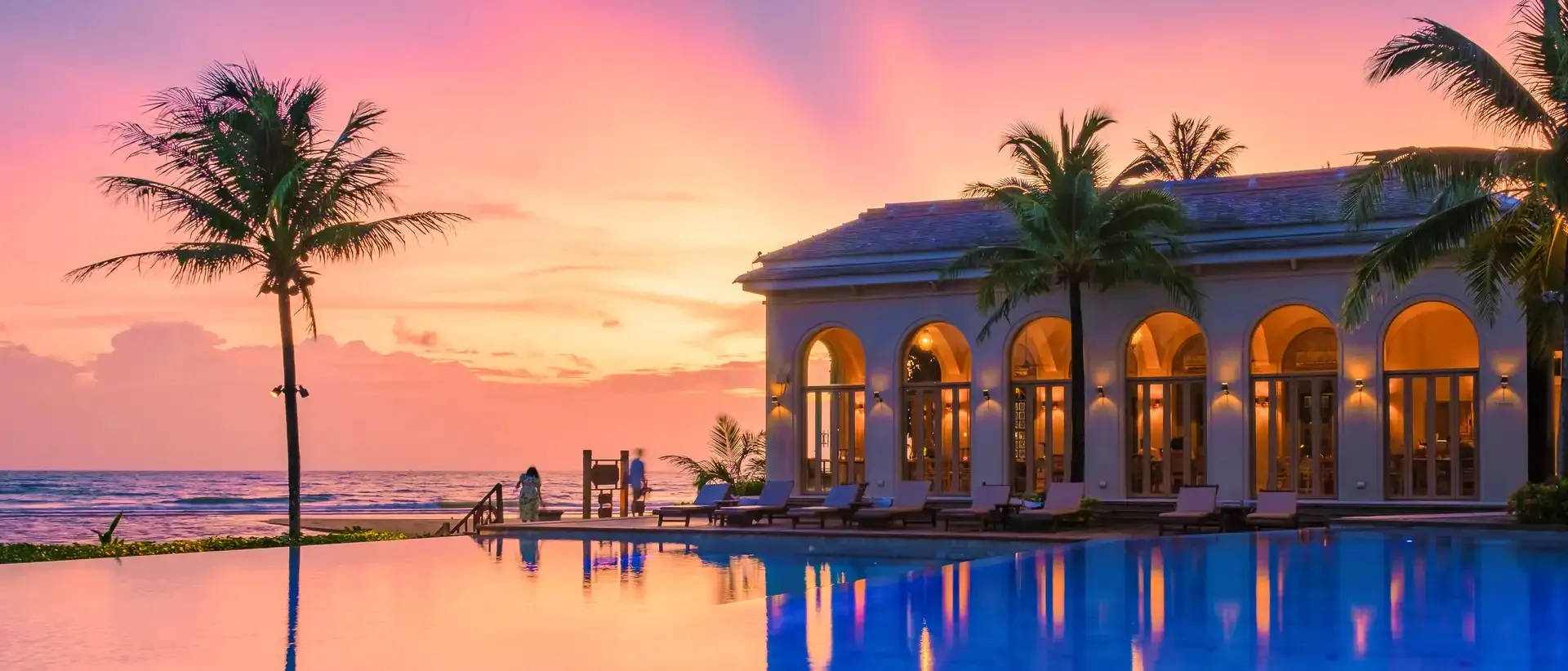 Resort pool and arcaded pavilion glowing under a pink sunset sky